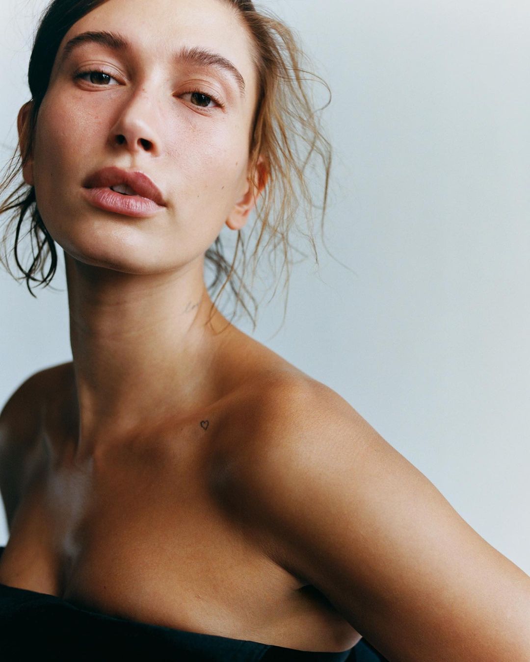 Portrait of an adult female with dark hair wearing a black top in a studio, fair skin, and a tiny heart tattoo on her shoulder.