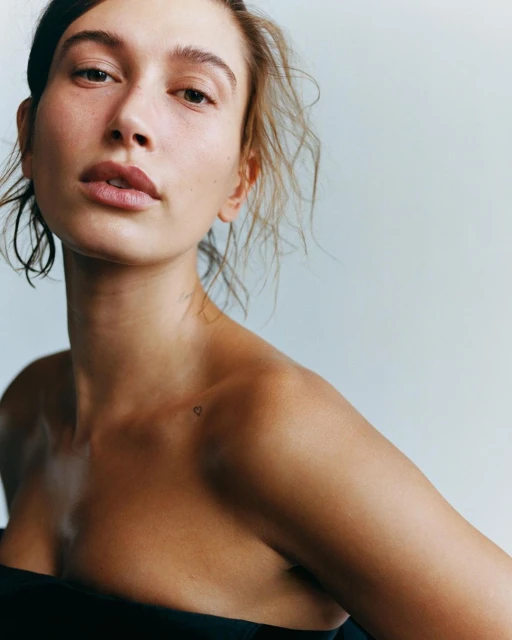 Portrait of an adult female with dark hair wearing a black top in a studio, fair skin, and a tiny heart tattoo on her shoulder.