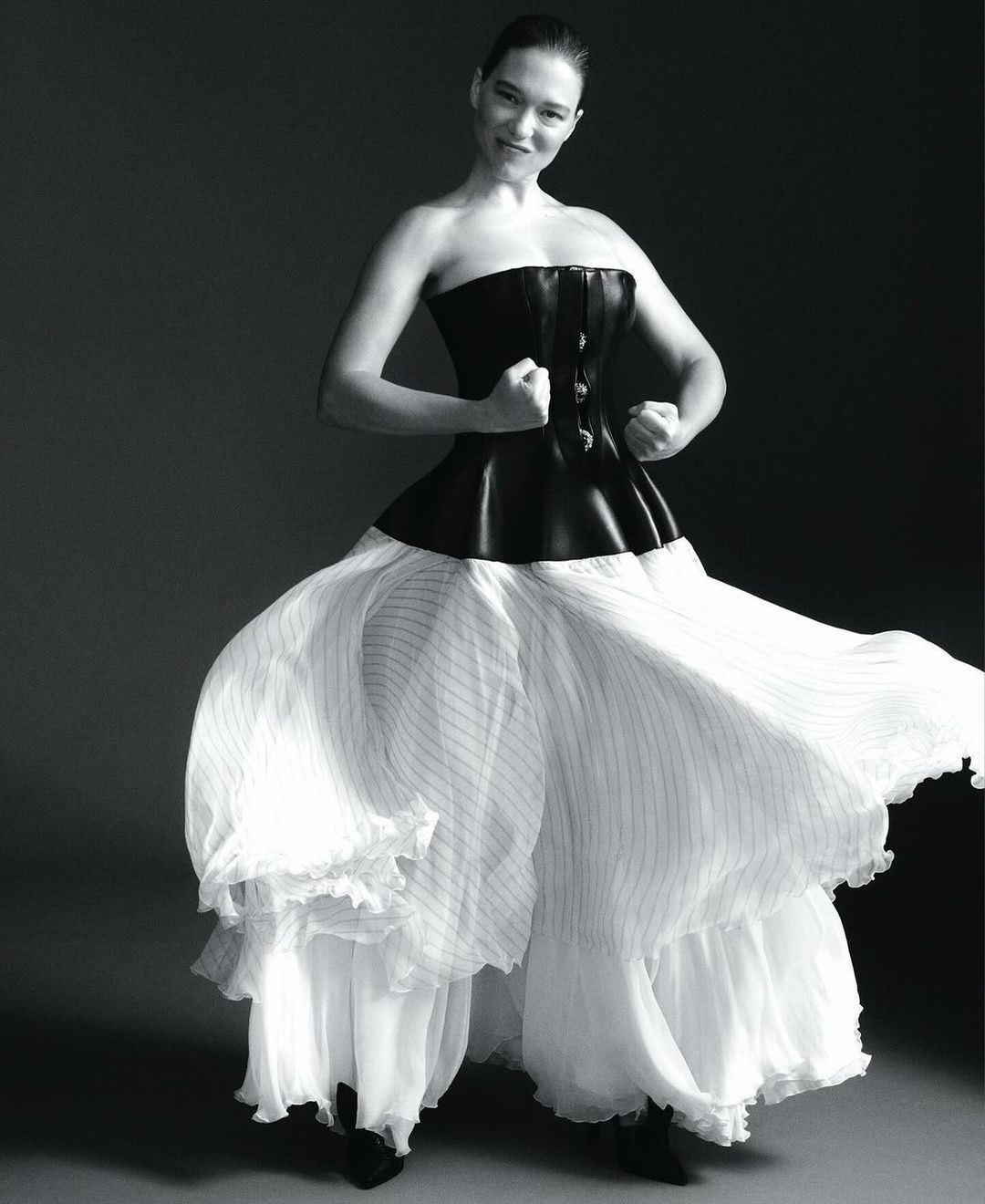 Black and white studio shot of an adult woman in a corset and billowing dress, arms raised, smiling against a gray backdrop.