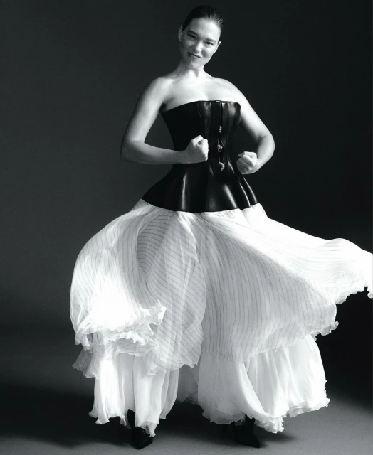 Black and white studio shot of an adult woman in a corset and billowing dress, arms raised, smiling against a gray backdrop.