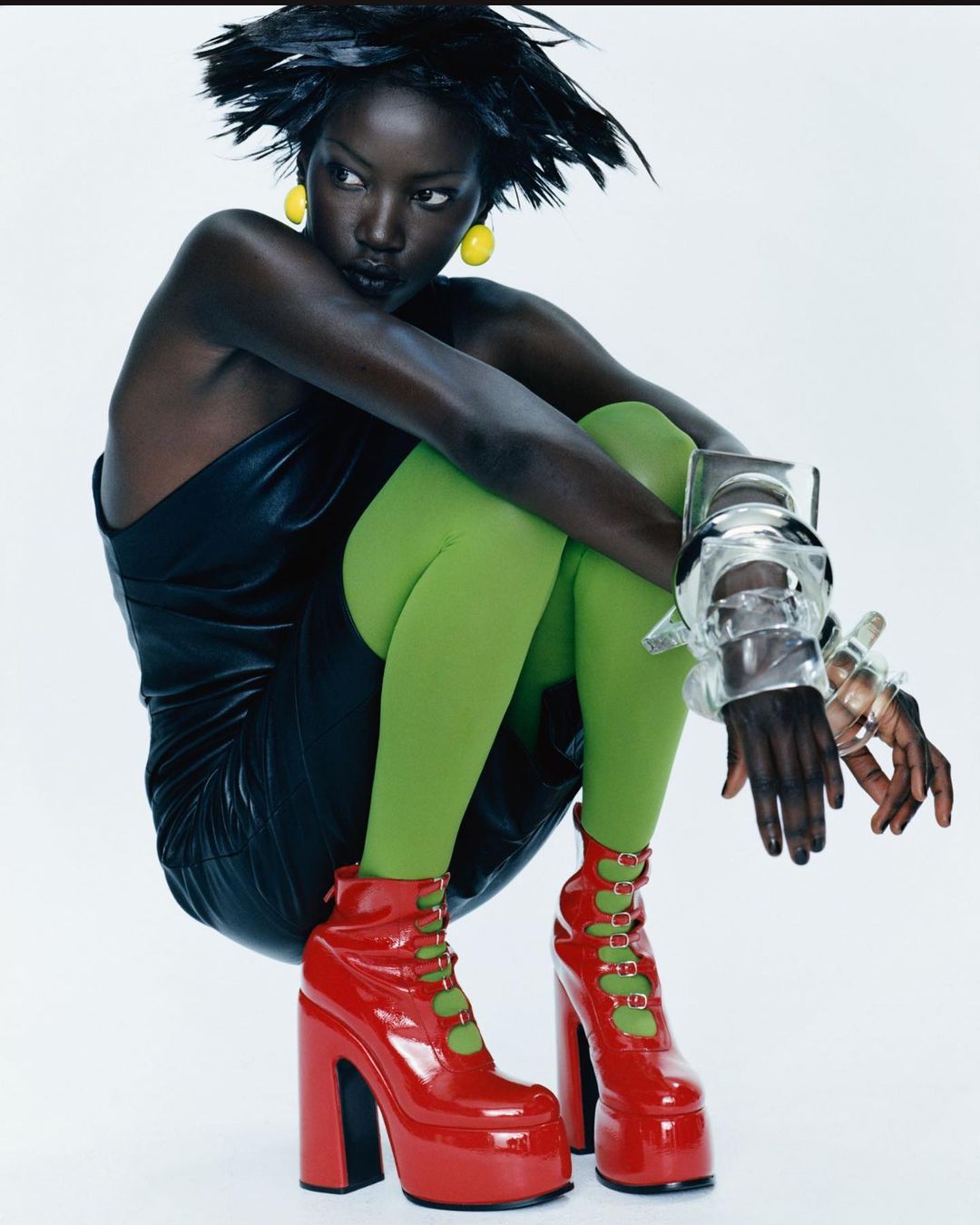 Fashionable dark-skinned woman in a black dress, green tights, and red platform boots poses in a studio with bright lighting.