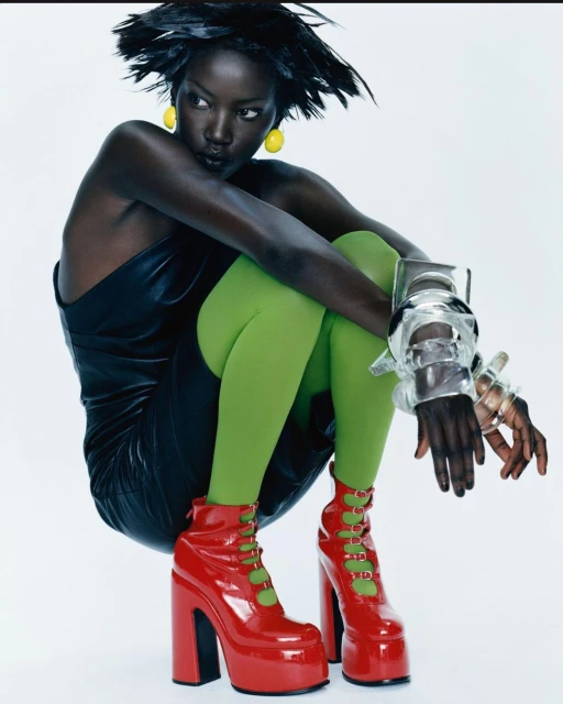 Fashionable dark-skinned woman in a black dress, green tights, and red platform boots poses in a studio with bright lighting.