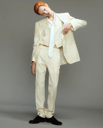 Model with red hair wearing a cream three-piece suit and white tie against a plain grey seamless background.