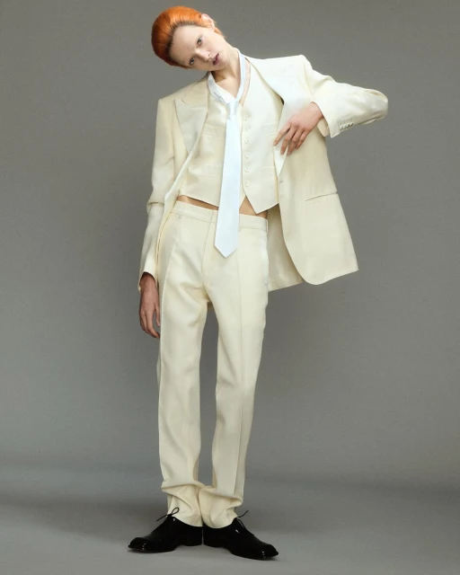 Androgynous fashion model poses in an off-white suit with a vest and tie, against a neutral grey background in a studio setting with black patent-leather shoes.