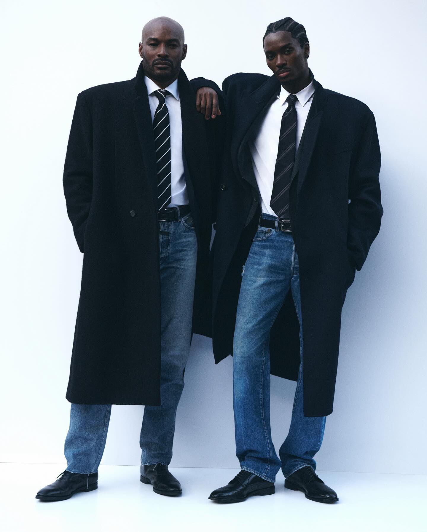 Two adult men in black overcoats and blue jeans stand close together indoors, exuding confidence and style in a fashion setting.