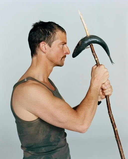 Profile shot of a man in a tank top holding a stick with a fish speared on it, set against a plain grey background in a photo studio.