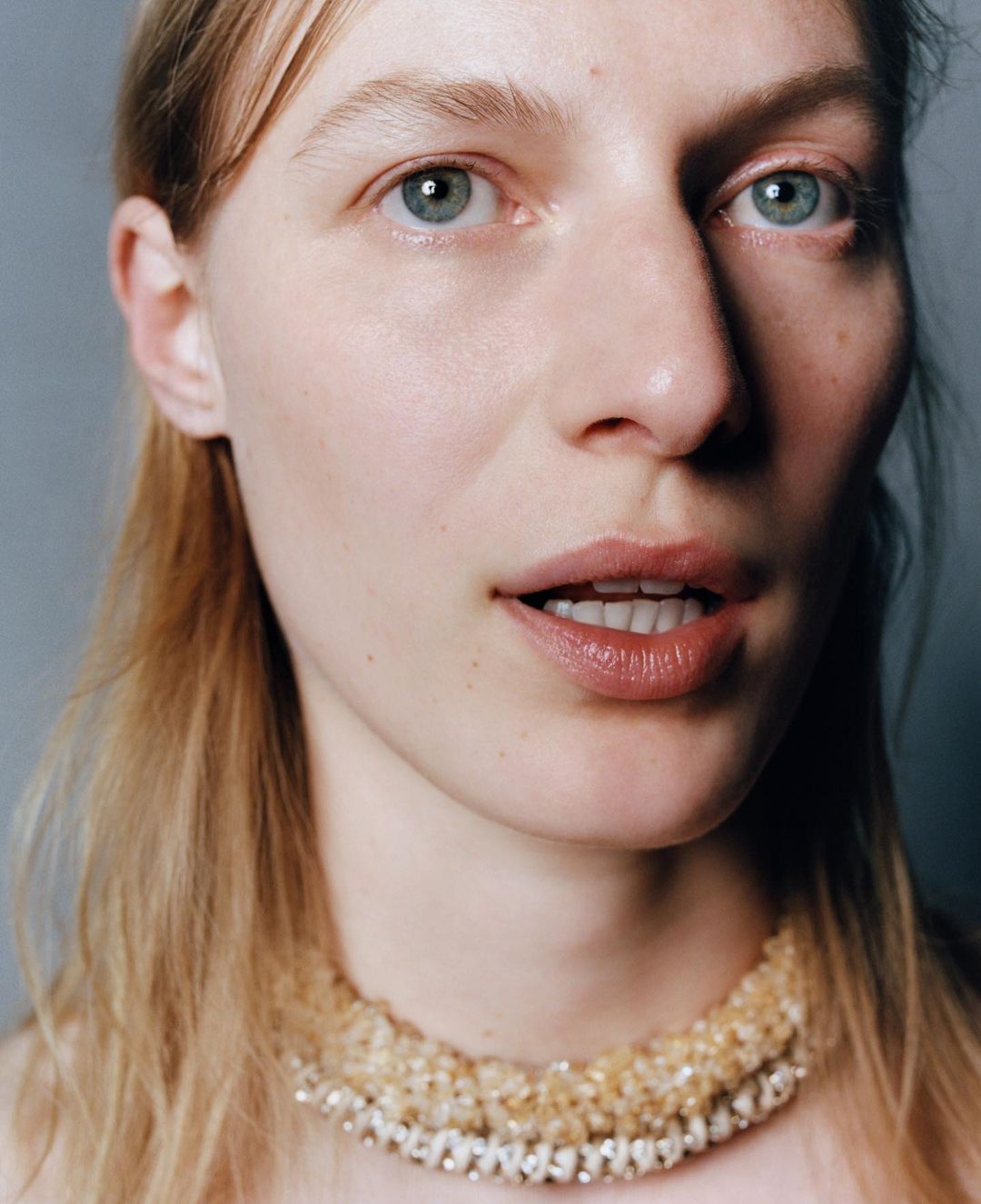 Close-up portrait of a fair-skinned adult woman with blue eyes and blonde hair wearing a beaded collar necklace against a gray background.