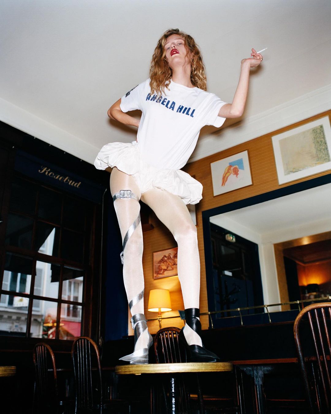 Fashionable woman in a white t-shirt and tutu stands on a table indoors holding a cigarette, showcasing edgy style and confidence.