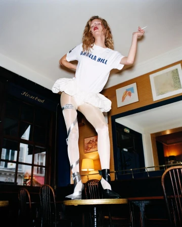 Fashionable woman in a white t-shirt and tutu stands on a table indoors holding a cigarette, showcasing edgy style and confidence.