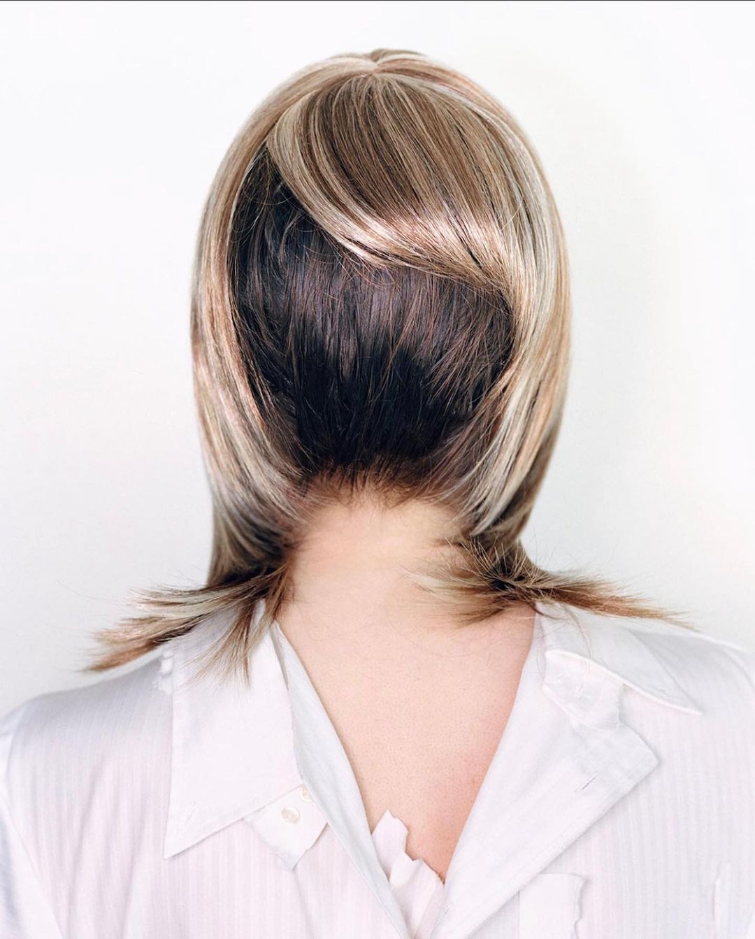 Close-up from the back of an adult female wearing a blond wig with dark roots and a white button-down shirt against a white background.