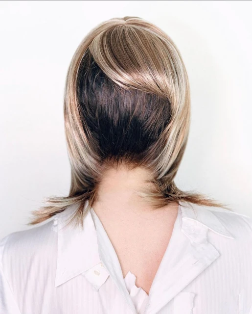 Close-up from the back of an adult female wearing a blond wig with dark roots and a white button-down shirt against a white background.