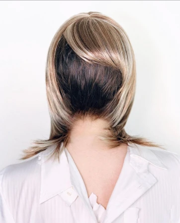 Close-up from the back of an adult female wearing a blond wig with dark roots and a white button-down shirt against a white background.