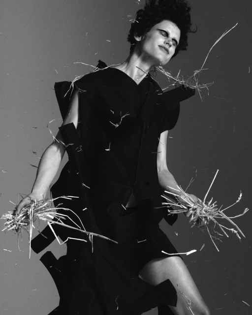 Monochrome fashion shot of a female model in a dark dress, holding straw bundles against a light gray backdrop, styled with dark eye makeup.