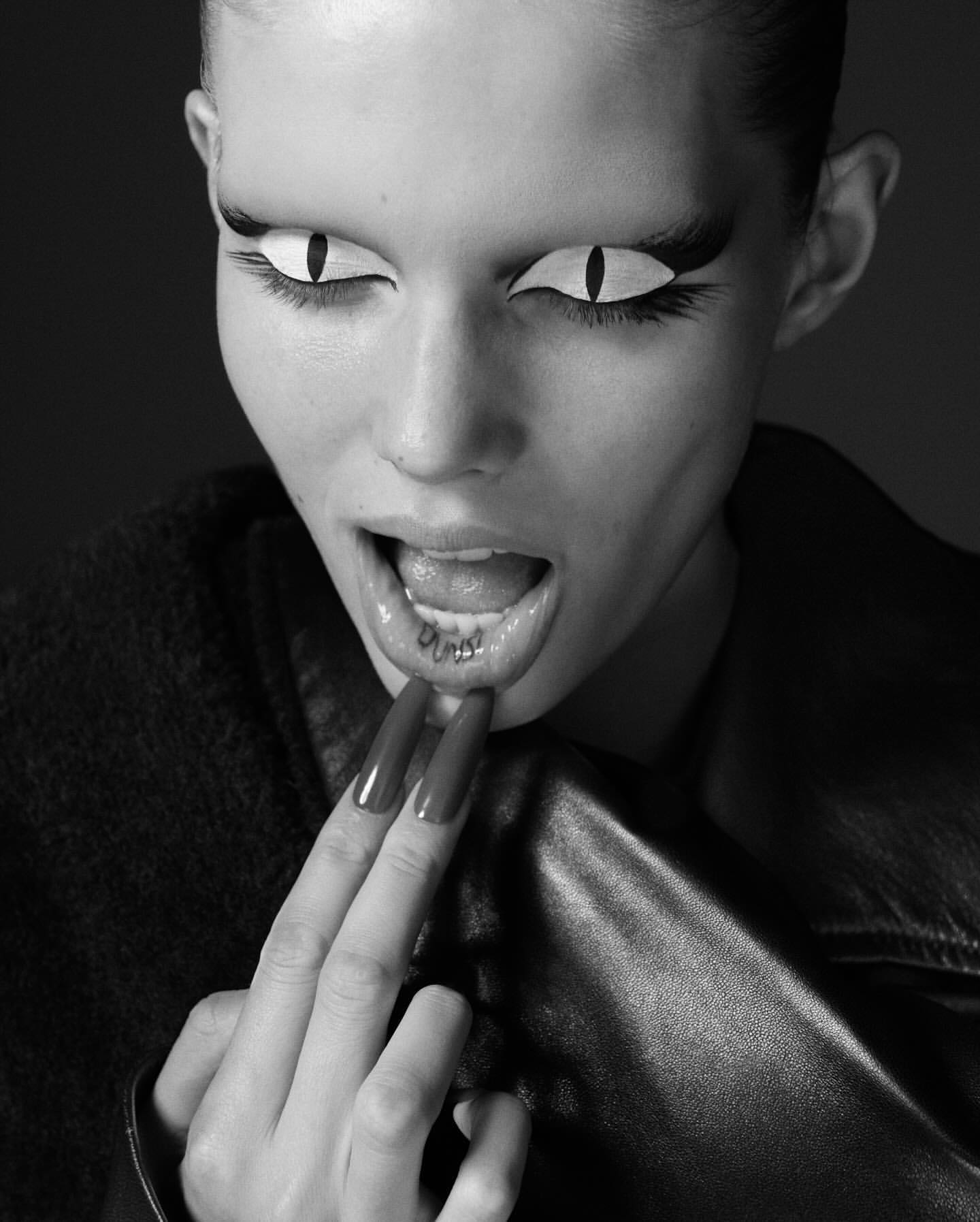 Monochrome close-up of a woman with bold, stylized eye makeup resembling feline eyes, a lip piercing, and long fingernails.