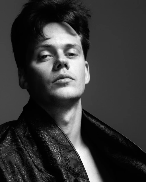 Black and white portrait of a young adult male wearing a patterned black robe against a grey background in a studio environment.