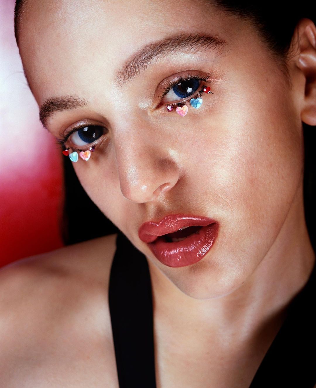 Close-up of a woman's face with heart-shaped decorations around her eyes, showcasing beauty and creative makeup with a red backdrop.