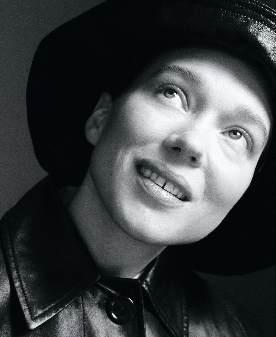 Monochrome portrait of a person with a hat and jacket looking up, set against a plain gray background in a studio environment.