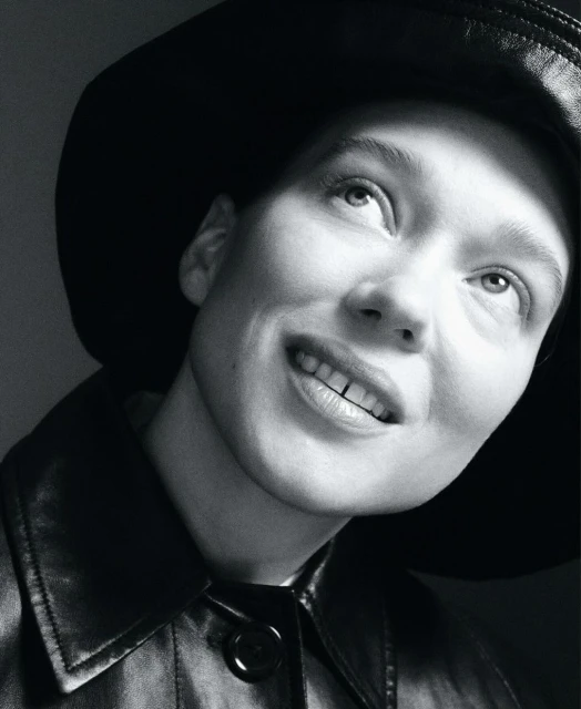 Monochrome portrait of a person with a hat and jacket looking up, set against a plain gray background in a studio environment.