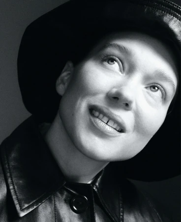Monochrome portrait of a person with a hat and jacket looking up, set against a plain gray background in a studio environment.
