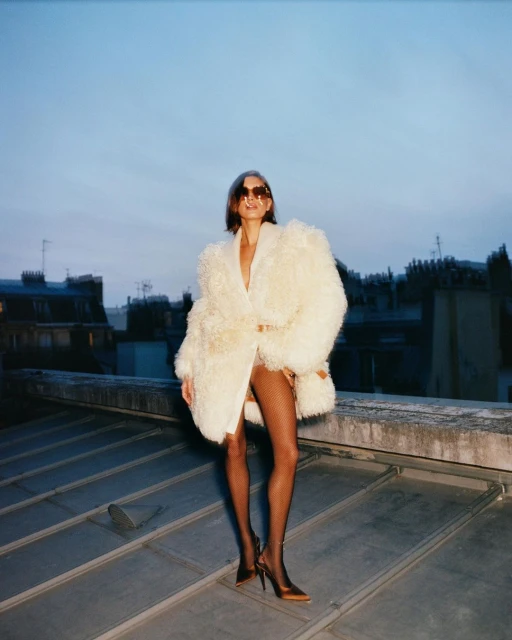 Elegant woman poses on rooftop wearing oversized white fur coat, black hose, heels, and sunglasses in an urban setting at dusk.