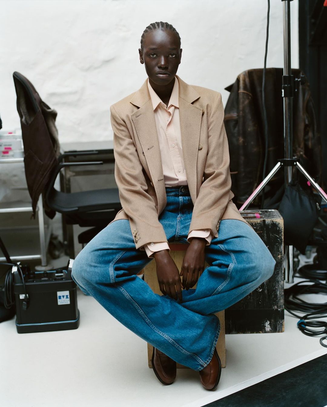 An adult Black female sits cross-legged on a wooden stool in a photo studio wearing a blazer, jeans, button-down shirt and brown shoes.