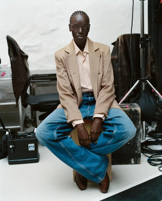 An adult Black female sits cross-legged on a wooden stool in a photo studio wearing a blazer, jeans, button-down shirt and brown shoes.