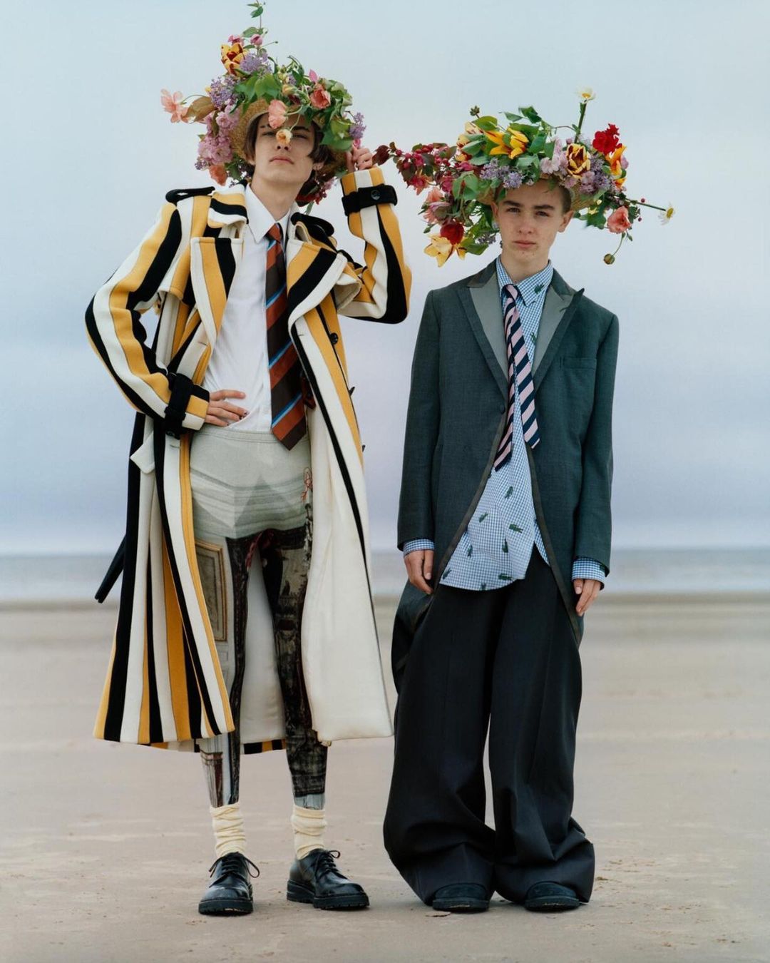 Two young men stand on a sandy beach wearing floral headpieces and avant-garde fashion ensembles in a creative outdoor editorial shoot.