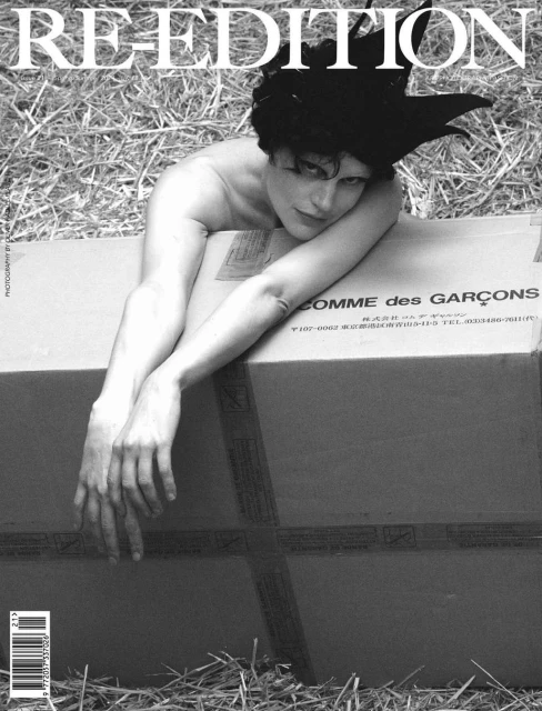 Black and white editorial fashion portrait of a bare-chested person with a large sculptural headpiece, lying on a cardboard box.