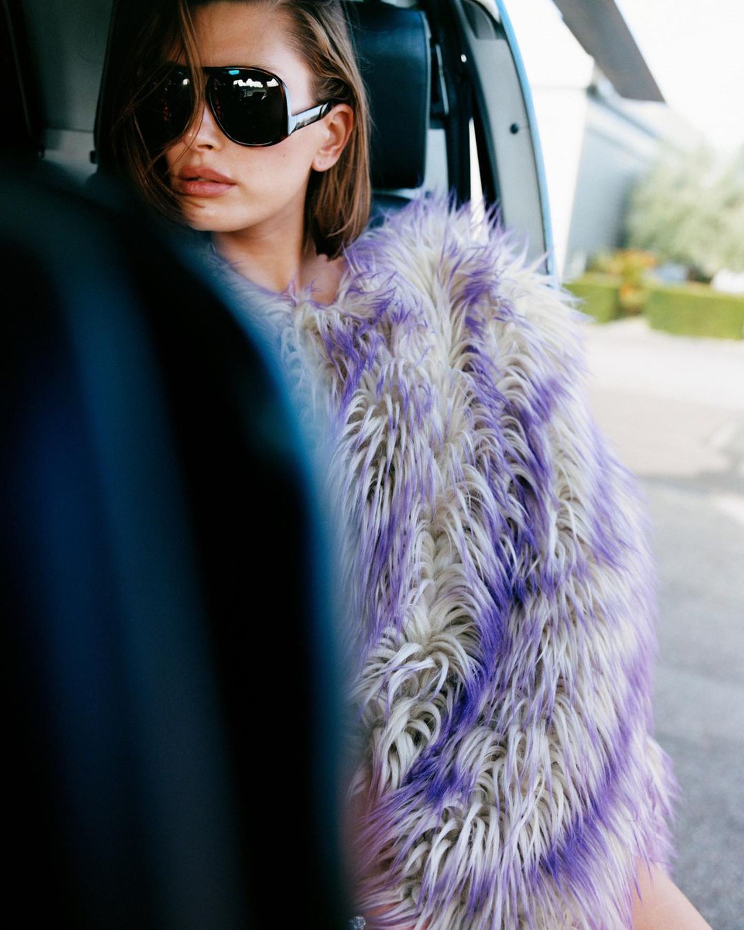 A woman wearing large black sunglasses and a fur vest sits in a helicopter with a green shrub in the background on a sunny day.
