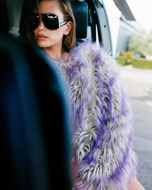A woman wearing large black sunglasses and a fur vest sits in a helicopter with a green shrub in the background on a sunny day.