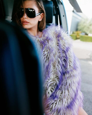 A woman wearing large black sunglasses and a fur vest sits in a helicopter with a green shrub in the background on a sunny day.