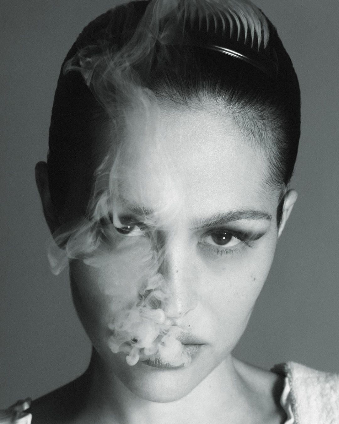 Monochrome portrait of a young adult woman with dramatic makeup posing with smoke in front of a neutral background in studio lighting.