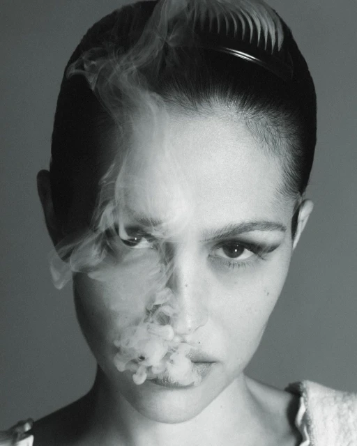 Monochrome portrait of a young adult woman with dramatic makeup posing with smoke in front of a neutral background in studio lighting.