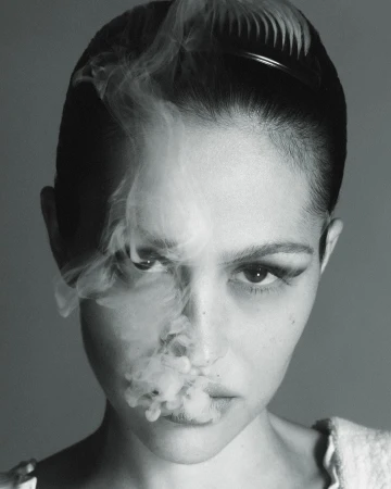 Monochrome portrait of a young adult woman with dramatic makeup posing with smoke in front of a neutral background in studio lighting.