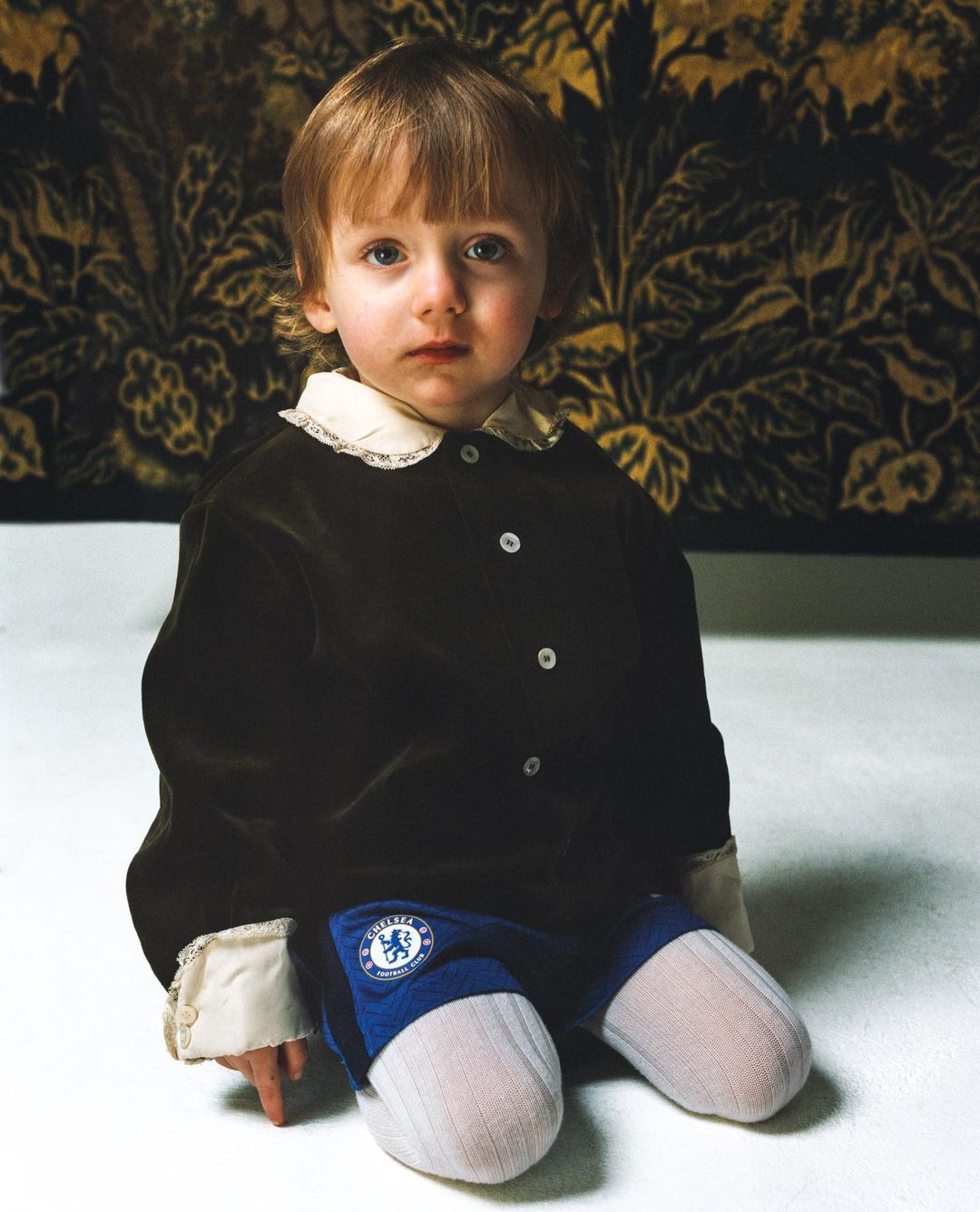 Portrait of a young child with blond hair, wearing a brown jacket and blue bottoms, seated on a white surface in front of a floral tapestry.