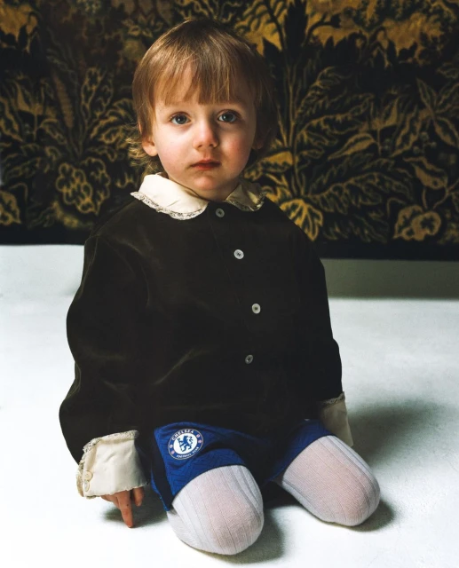 Portrait of a young child with blond hair, wearing a brown jacket and blue bottoms, seated on a white surface in front of a floral tapestry.