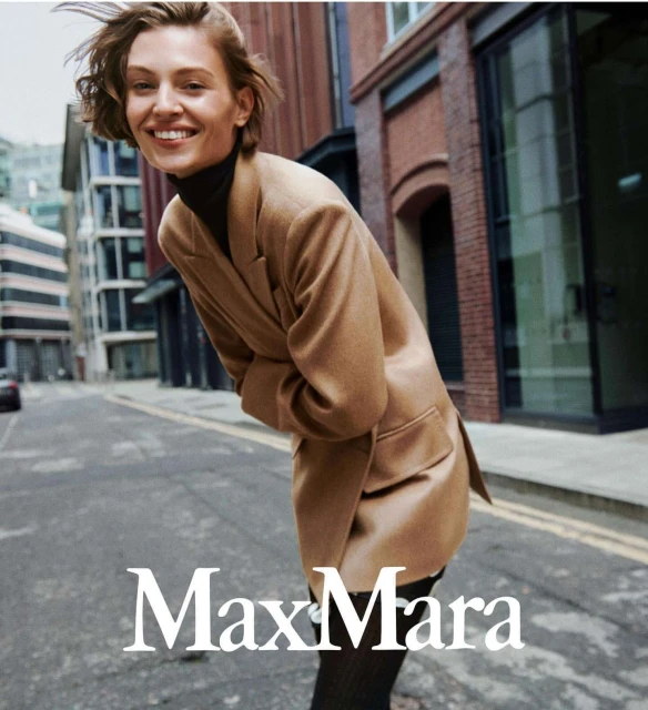Blonde woman in a camel-colored blazer smiles in an outdoor street scene. The Max Mara logo appears at the bottom.