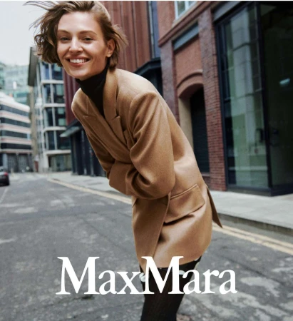 Blonde woman in a camel-colored blazer smiles in an outdoor street scene. The Max Mara logo appears at the bottom.