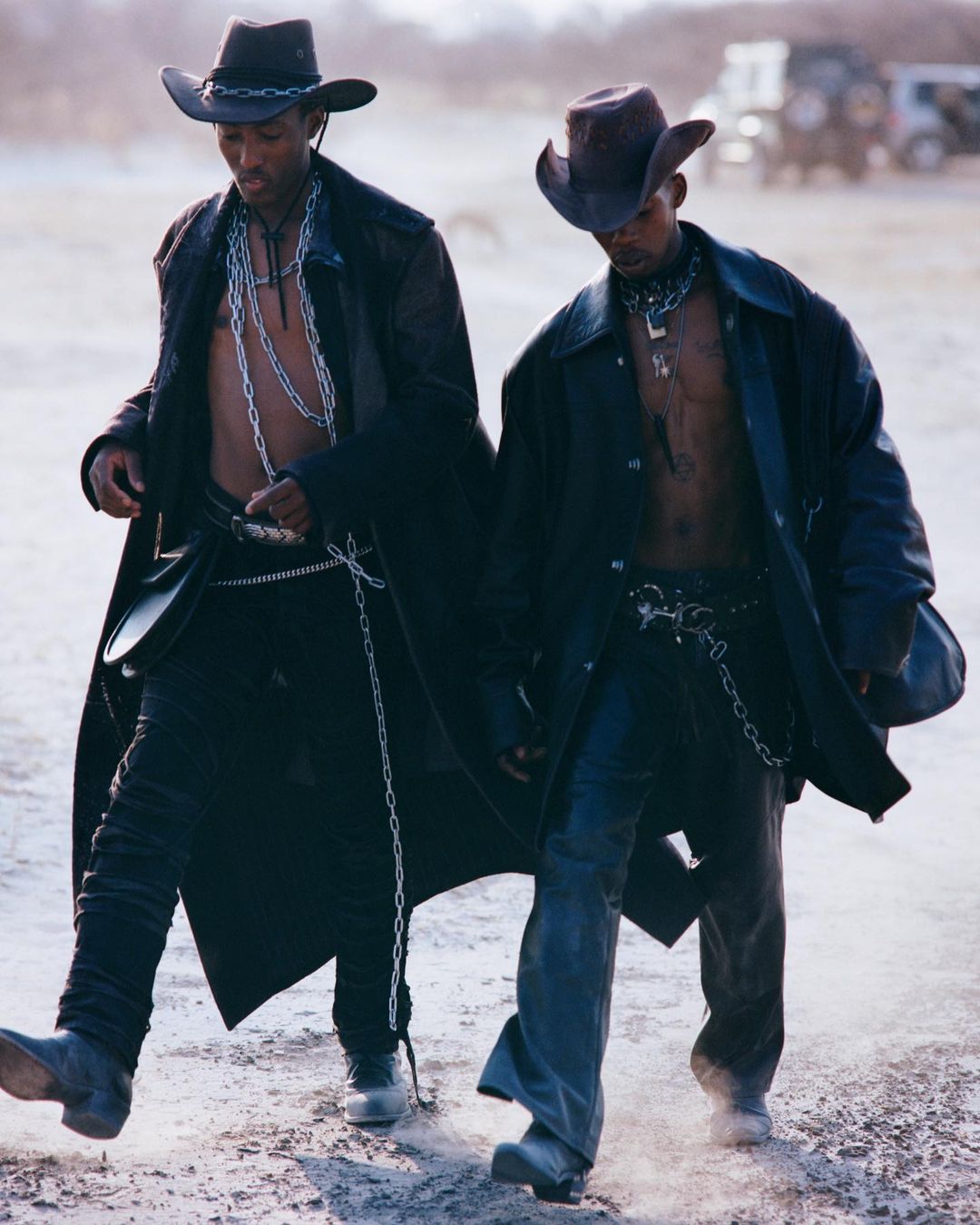 Two African American men in cowboy hats and long coats walk outdoors in a desert landscape with a safari vehicle in the background.