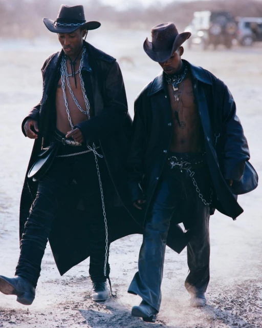 Two African American men in cowboy hats and long coats walk outdoors in a desert landscape with a safari vehicle in the background.