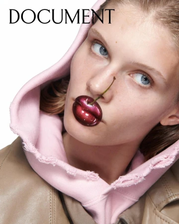 Close-up portrait of a woman wearing a pink hoodie holding a cherry between her lips by photographer Oliver Hadlee Pearch.