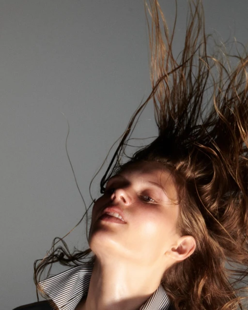 Woman with hair in motion, wearing a striped top, posing against a gray backdrop with natural light highlighting her face and shoulder.