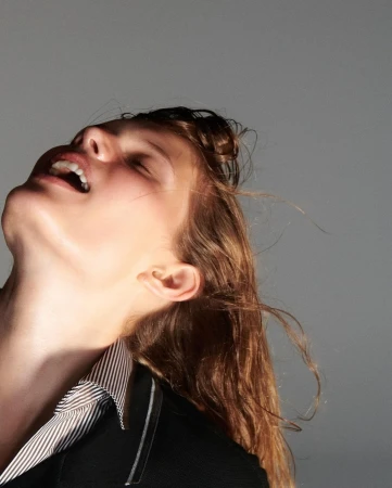 A woman with long brown hair smiles and looks up into the light, wearing a black blazer and striped shirt against a grey background.