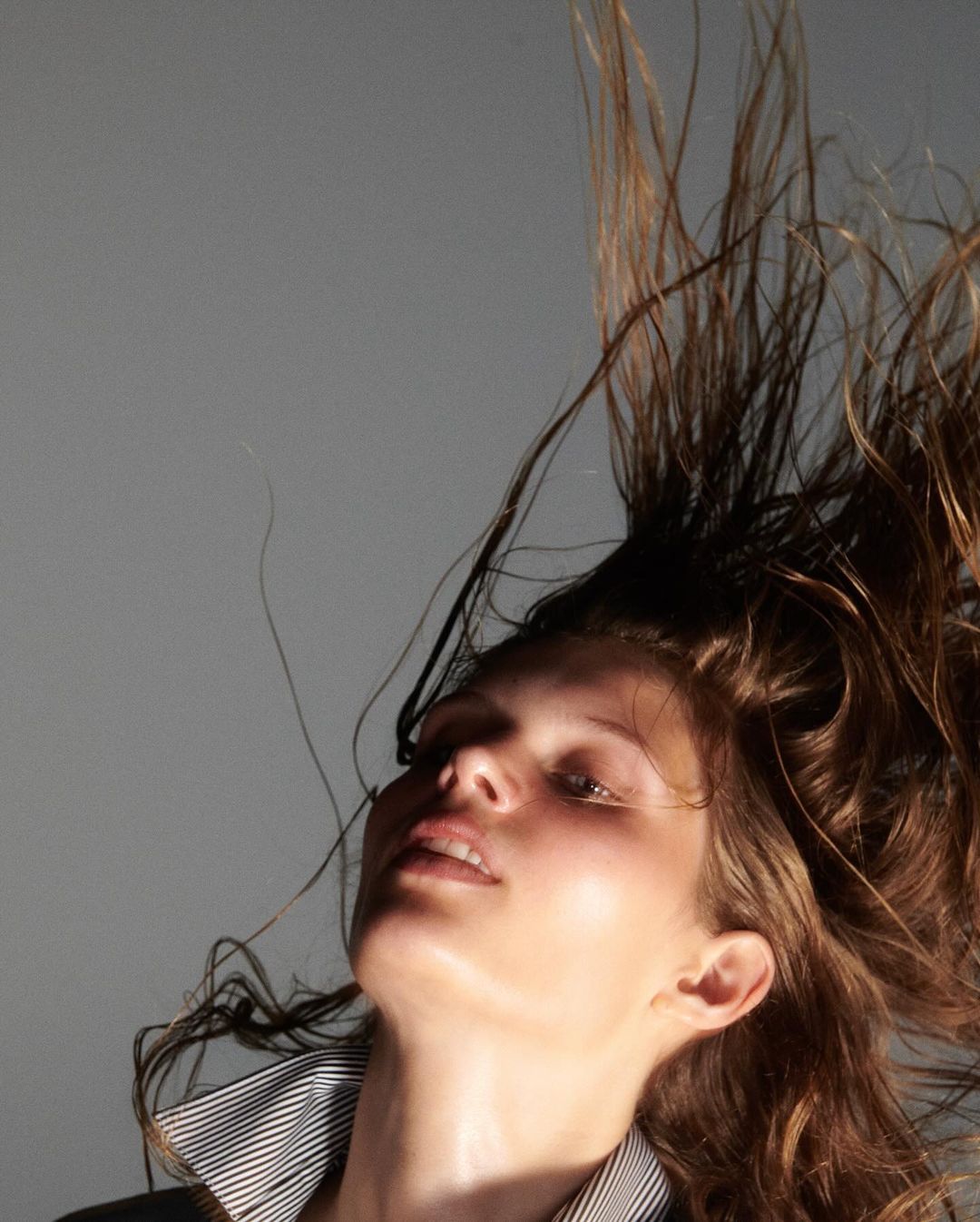 Low-angle shot of a woman with long brown hair, wearing a striped top, with her hair tossed upward against a grey studio backdrop.