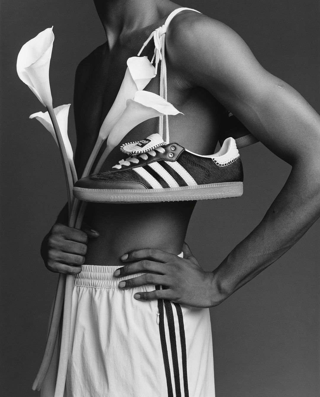 Monochrome studio shot of a dark-skinned shirtless adult male in shorts holding calla lilies, with an Adidas Samba shoe on his shoulder.