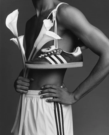 Monochrome studio shot of a dark-skinned shirtless adult male in shorts holding calla lilies, with an Adidas Samba shoe on his shoulder.
