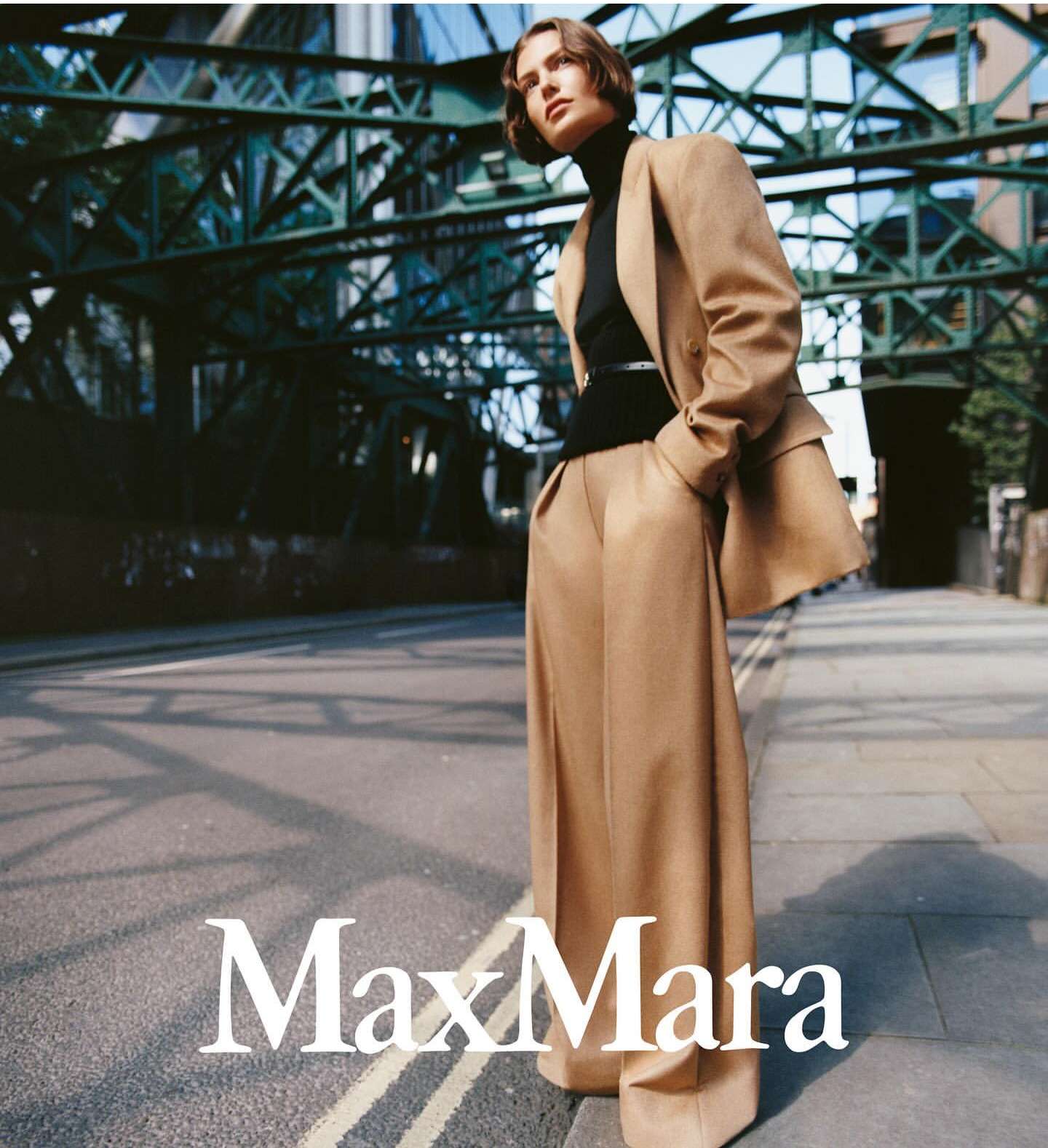 Woman in a camel-colored suit stands on a city street, posing in a fashion editorial with an overpass structure overhead.