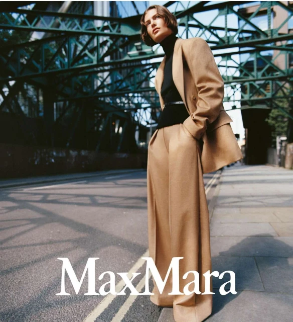 Woman in a camel-colored suit stands on a city street, posing in a fashion editorial with an overpass structure overhead.