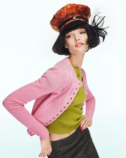 A fashion model poses in a pink cardigan, green top, and cap with feathers, against a bright white background in a studio setting.