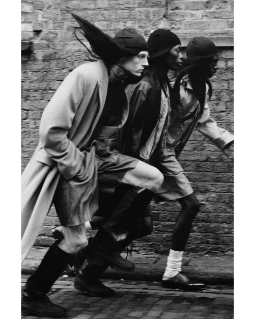 Three fashion models in hats and casual outerwear walk down a brick-paved street. Black and white editorial photography.